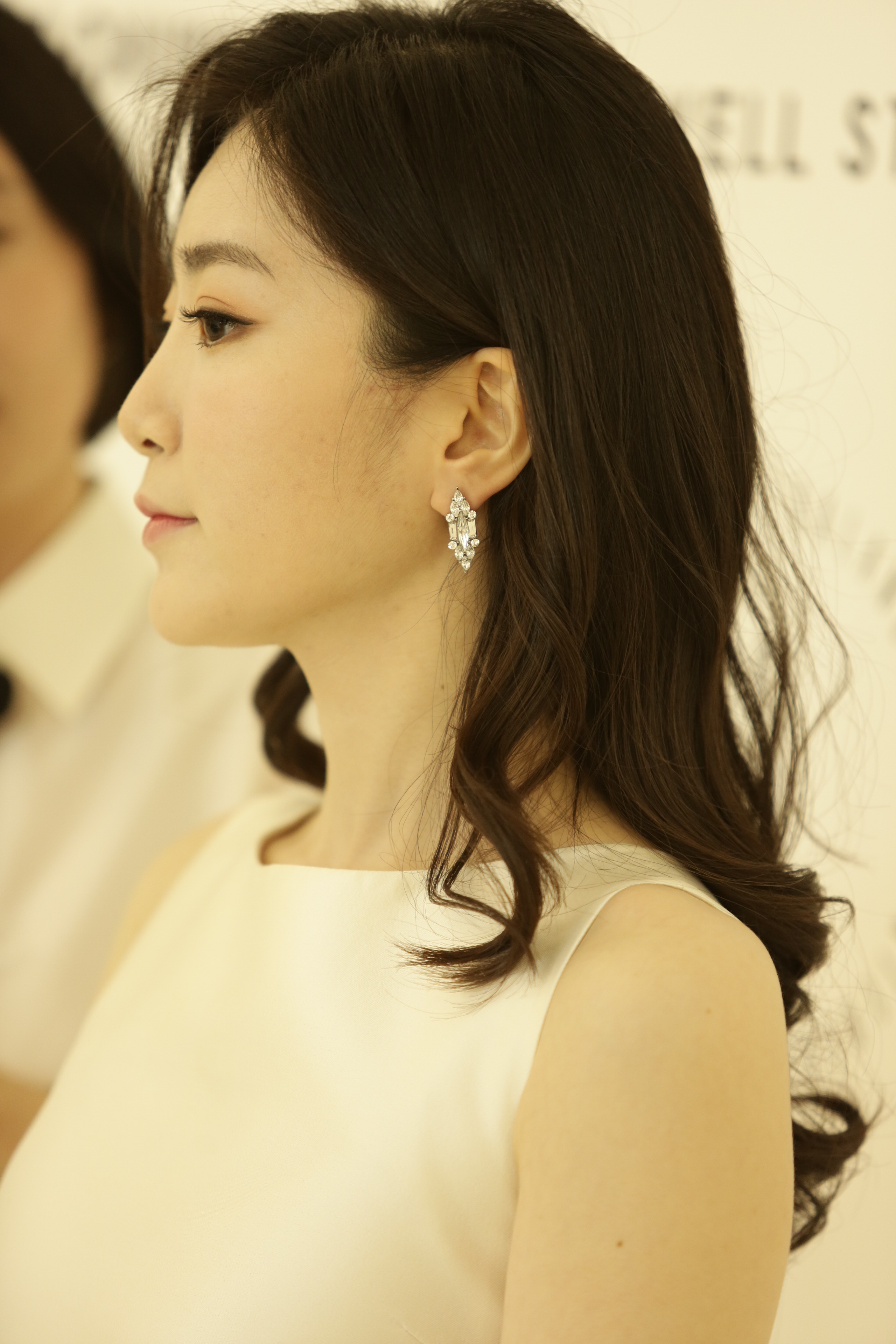 Woman in a white sleeveless dress with wavy dark hair, wearing Korean crystal earrings at a formal event