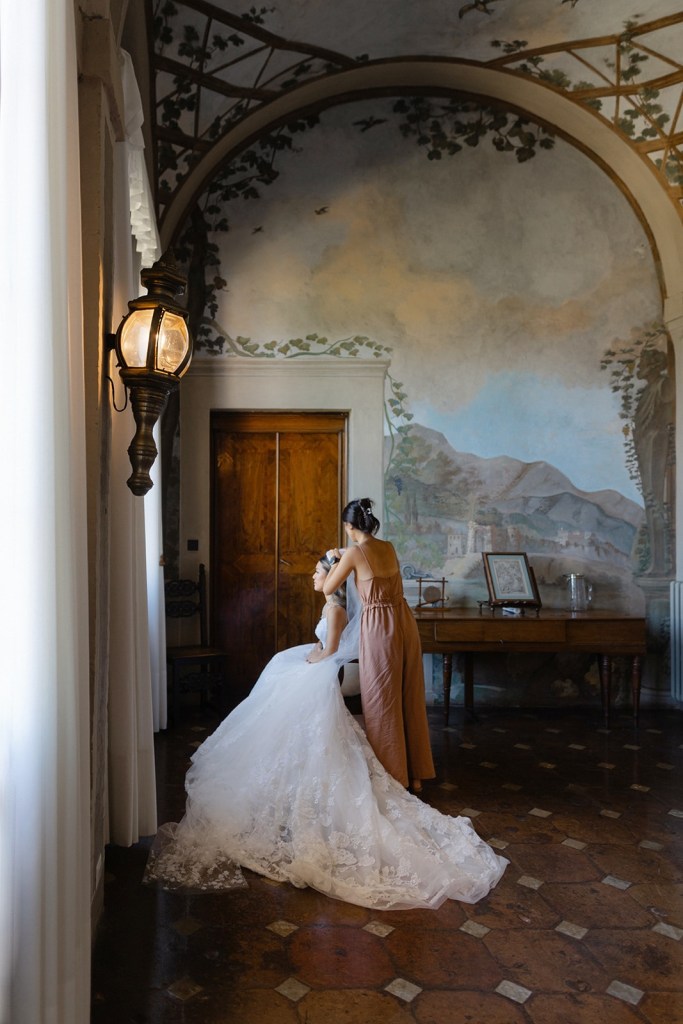 A bride in a stunning lace wedding dress is being assisted by a makeup artist in a beautifully decorated room with mural paintings and warm lighting.