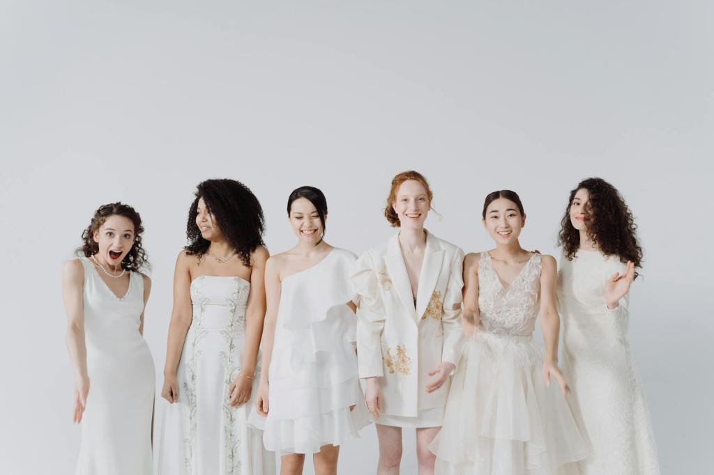 A diverse group of six women wearing various wedding dresses, showcasing different styles, fabrics and silhouettes, standing together against a neutral background.