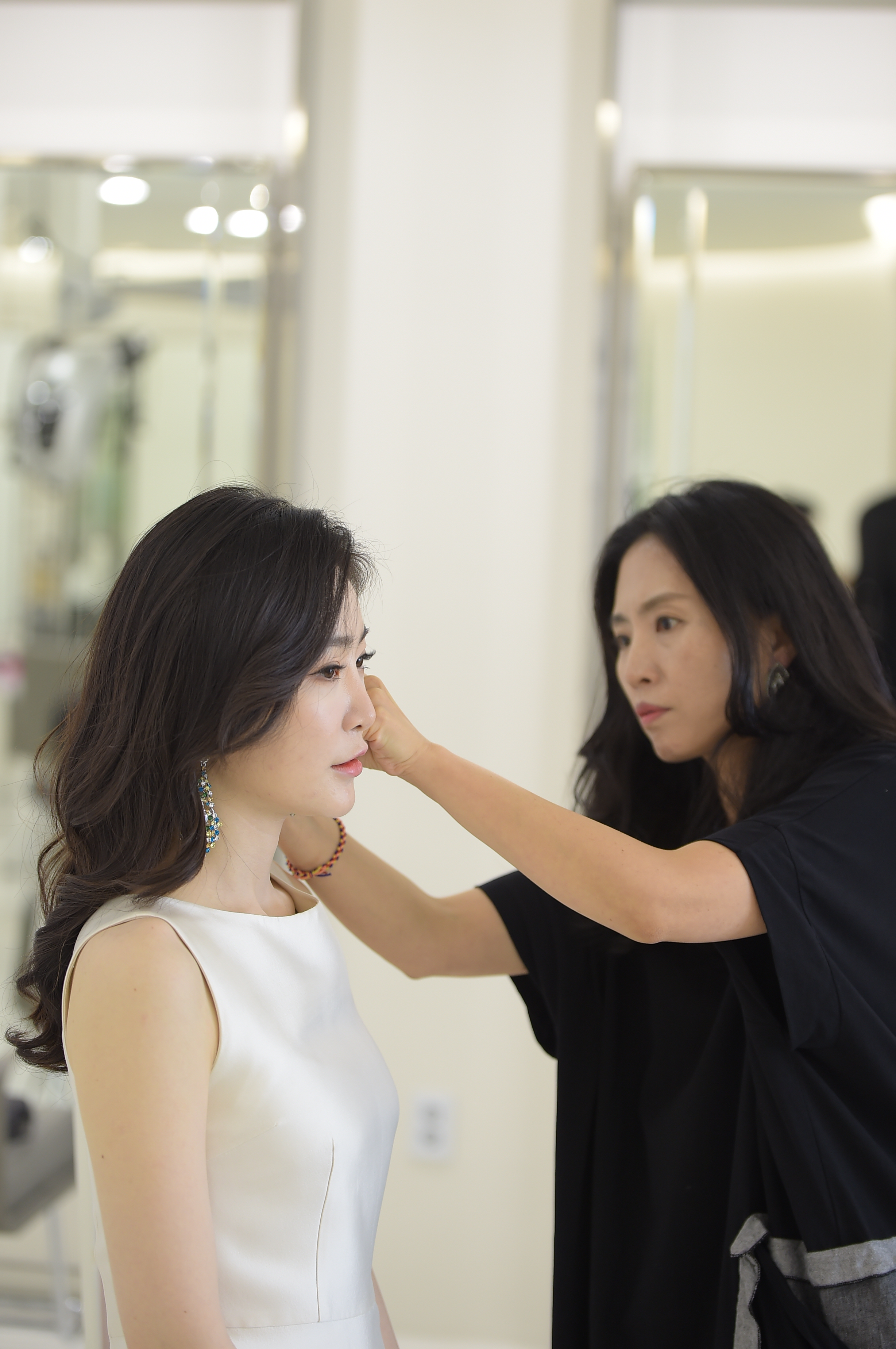 A stylist is adjusting the hair of a bride-to-be, who is wearing a white dress and an elegant earring, in a bright salon setting.