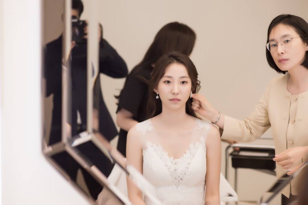 A bride in a wedding dress is being styled by a makeup artist in front of a mirror, showcasing elegance and focus during a bridal preparation session.